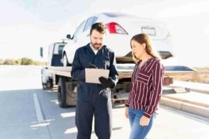 Tow truck operator reviewing paperwork with a driver beside a car loaded on a flatbed tow truck