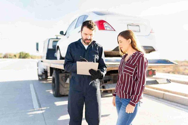 Tow truck operator reviewing paperwork with a driver beside a car loaded on a flatbed tow truck