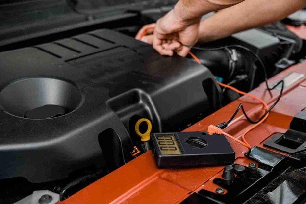 Digital multimeter resting on a car engine cover while a hand connects a cable to the battery