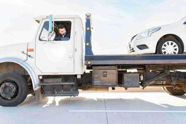 Flatbed tow truck transporting a vehicle with the driver visible in the cab