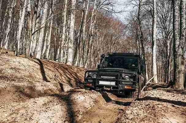 Black off-road vehicle driving through a muddy forest trail with deep ruts and puddles