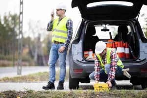 Two roadside workers in safety vests and hard hats placing equipment near an open vehicle trunk