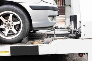 Front wheel of a silver car secured on a flatbed tow truck with a winch cable attached underneath